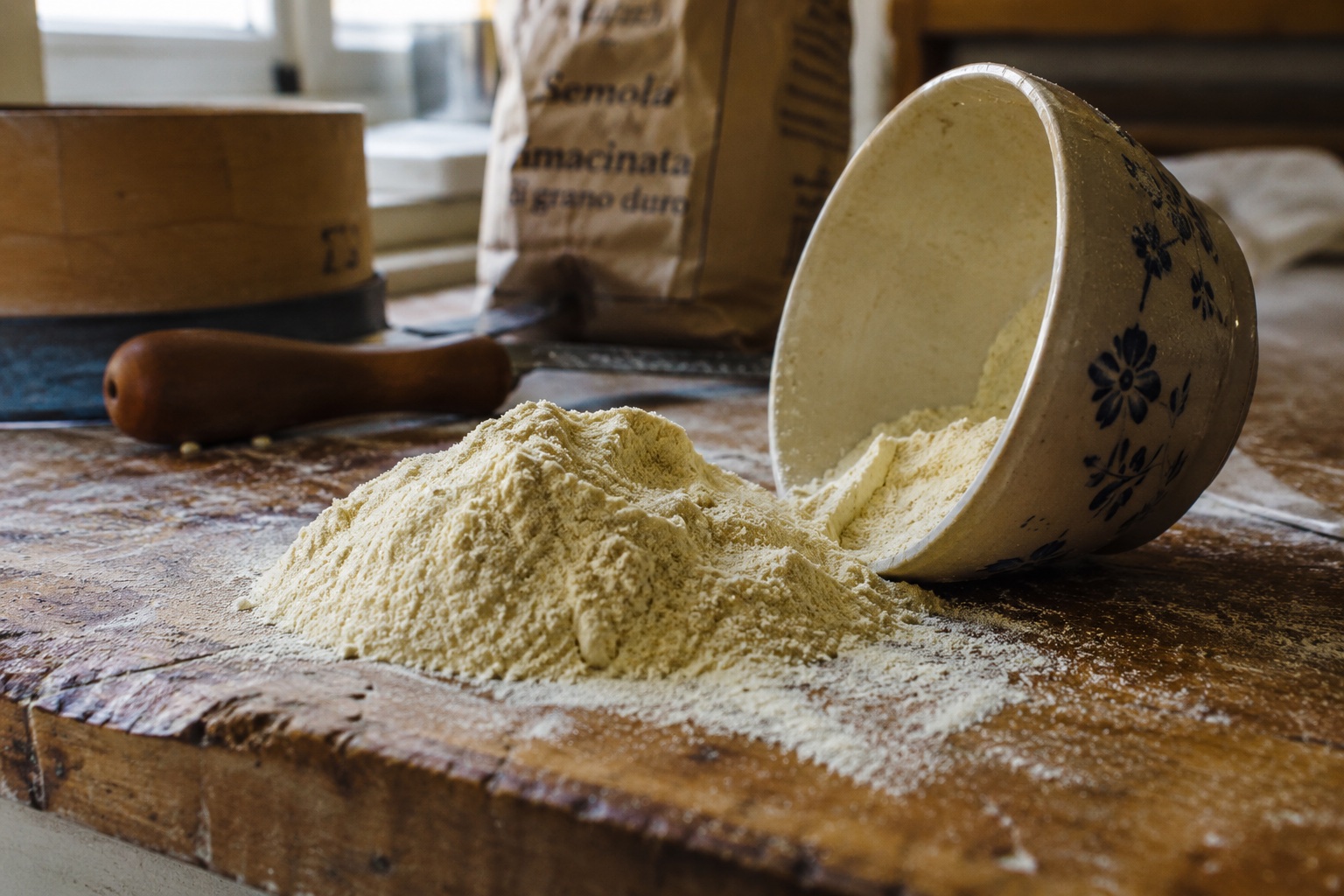 Durum semolina brašno se sipa iz keramične činije na drvenu radnu površinu, vreća "Semola Rimacinata di Grano Duro" u pozadini